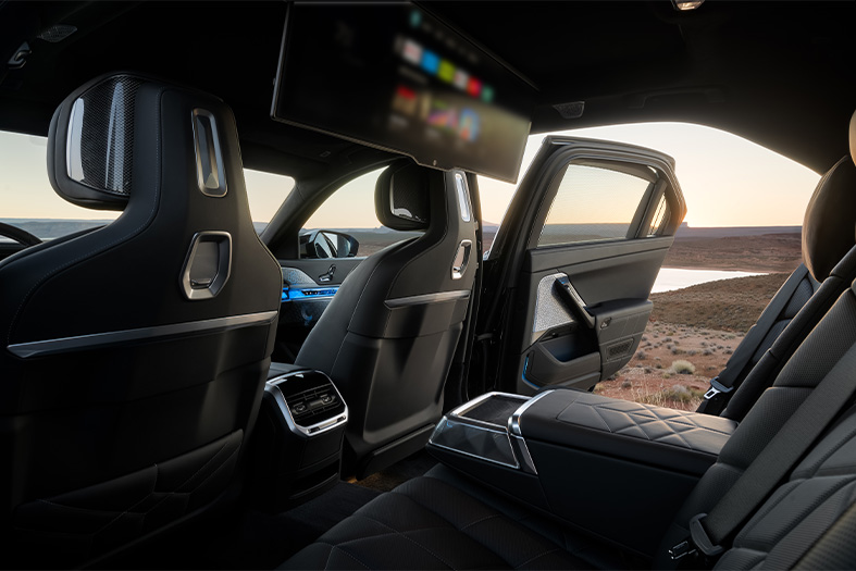  Backseat and theatre screen display of a 2026 BMW i7 M70 parked in flat, desert landscape with the sun setting in the background.
