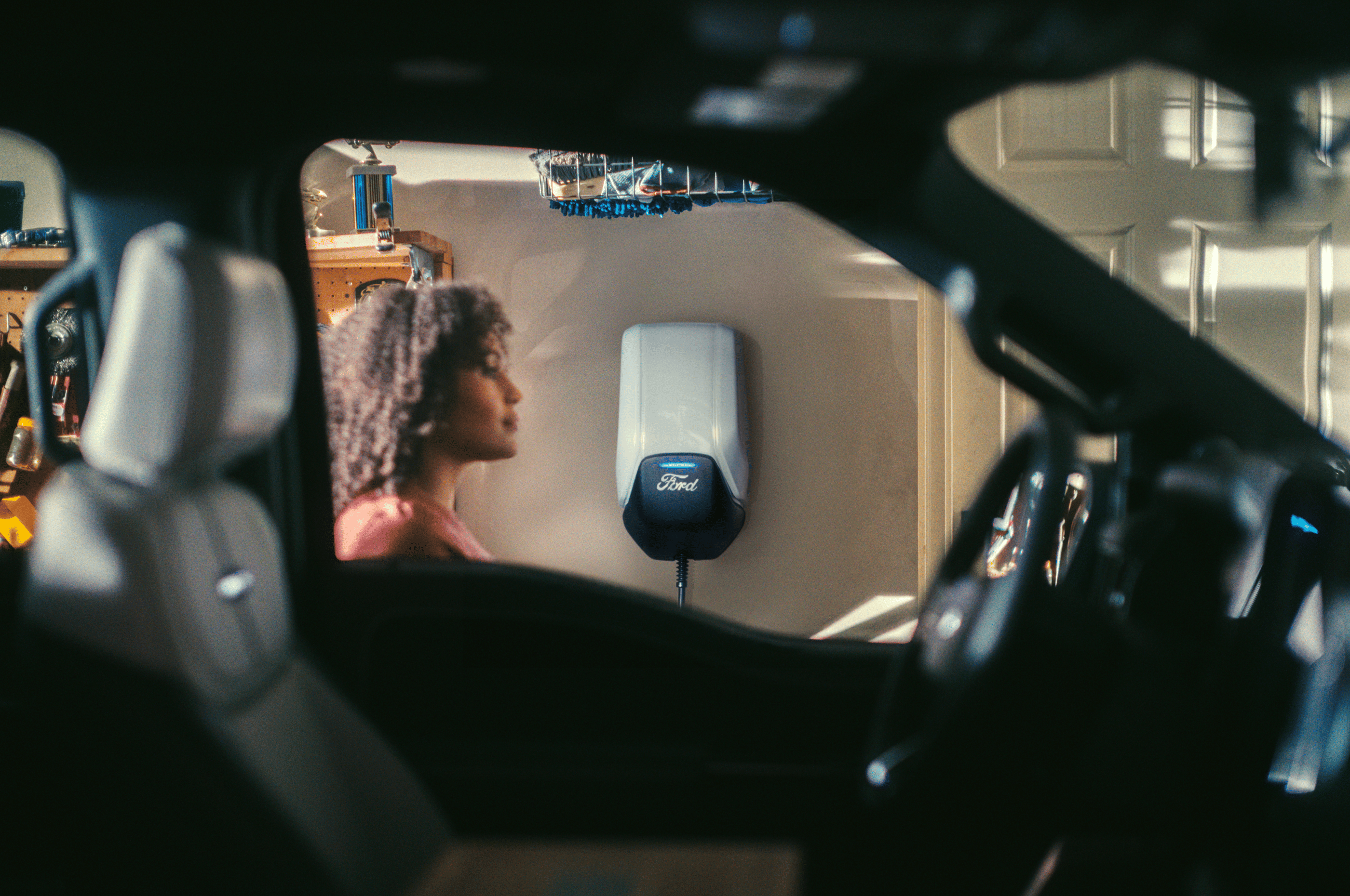 A Ford Connected Charge Station mounted on a garage wall with a woman seen through a Mustang Mach-e window.