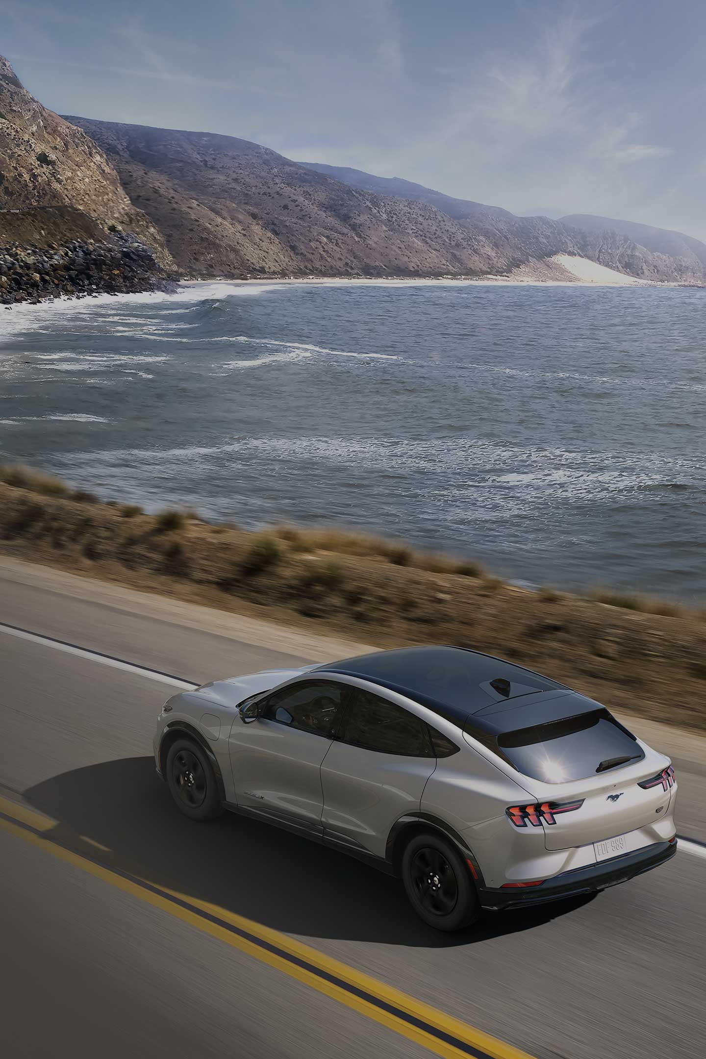 A Silver 2023 Ford Mustang Mach-E driving down the road along the ocean.