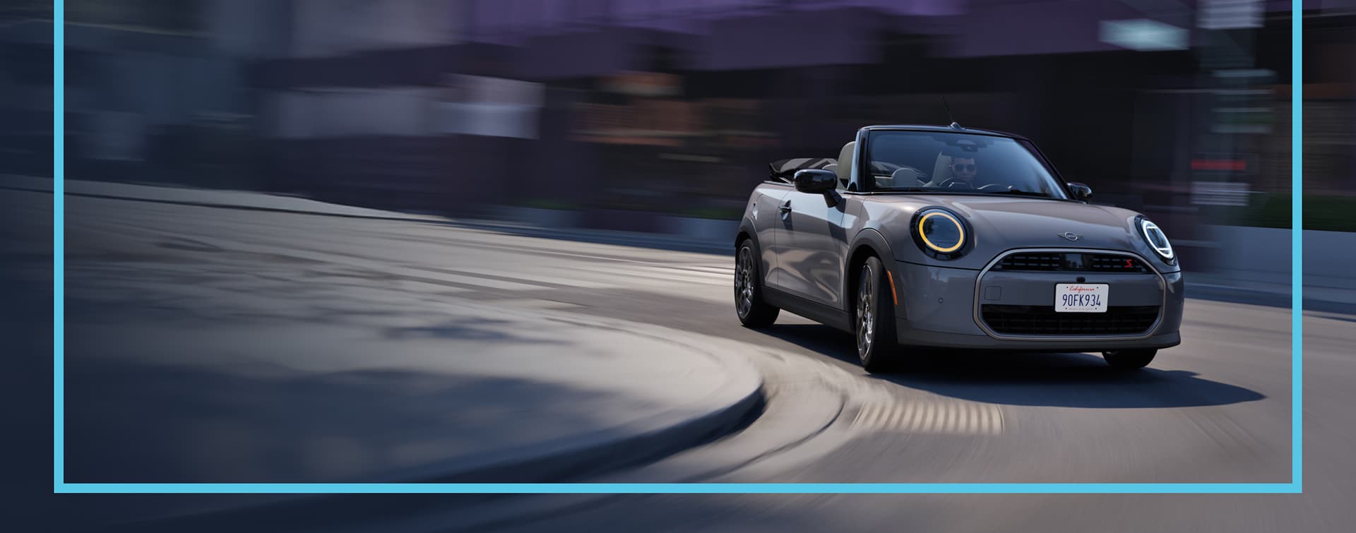Front view of a MINI Cooper Convertible driving on a city street with a blurred background.