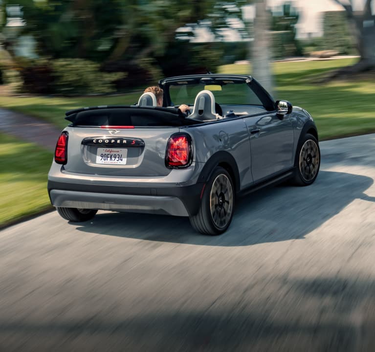 Rear-view of a MINI Cooper S Convertible in
        Icy Sunshine Blue turning around a bend in the road
        with a blurred, blue building in the background.