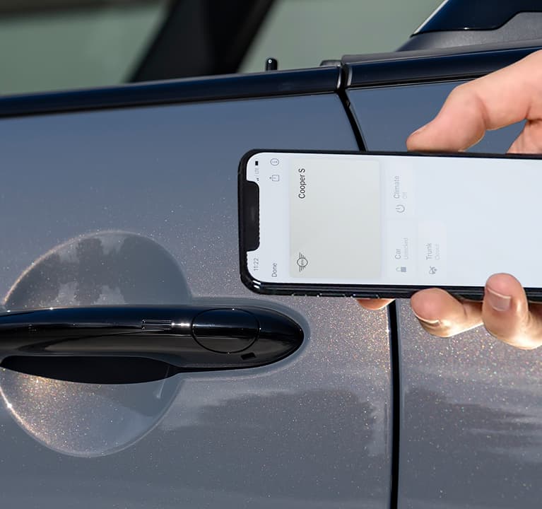 A hand holding up a smartphone open to the
        MINI App with a steering wheel in the background.