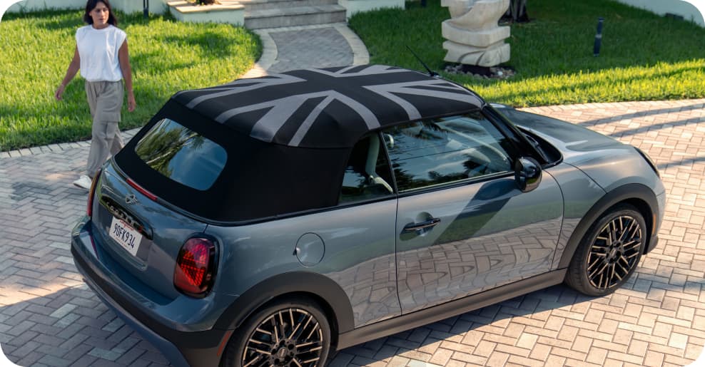 Woman standing behind the side view of grey MINI Cooper Convertible on a suburban street.