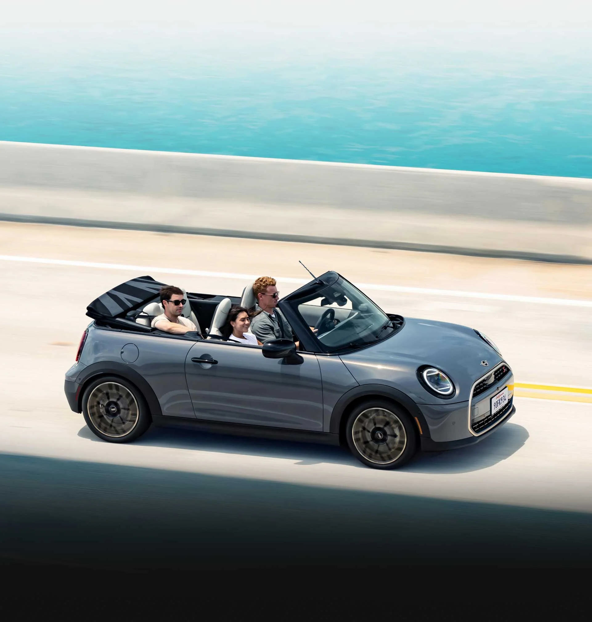 Grey MINI Cooper Convertible with three passengers driving on a seaside highway.