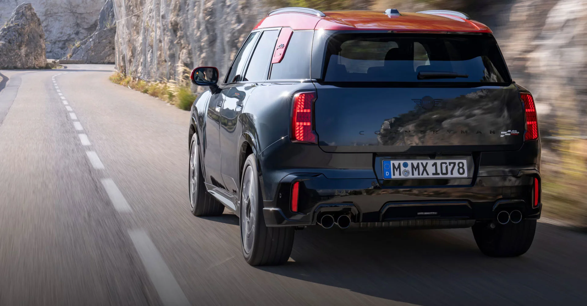 Angled rear-to-front view of the MINI JCW Countryman ALL4 driving down a road surrounded by a wall of rock and foliage.