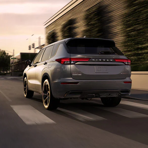 2026 Nissan Rogue Plug-in Hybrid rear view driving on city street approaching sunset.