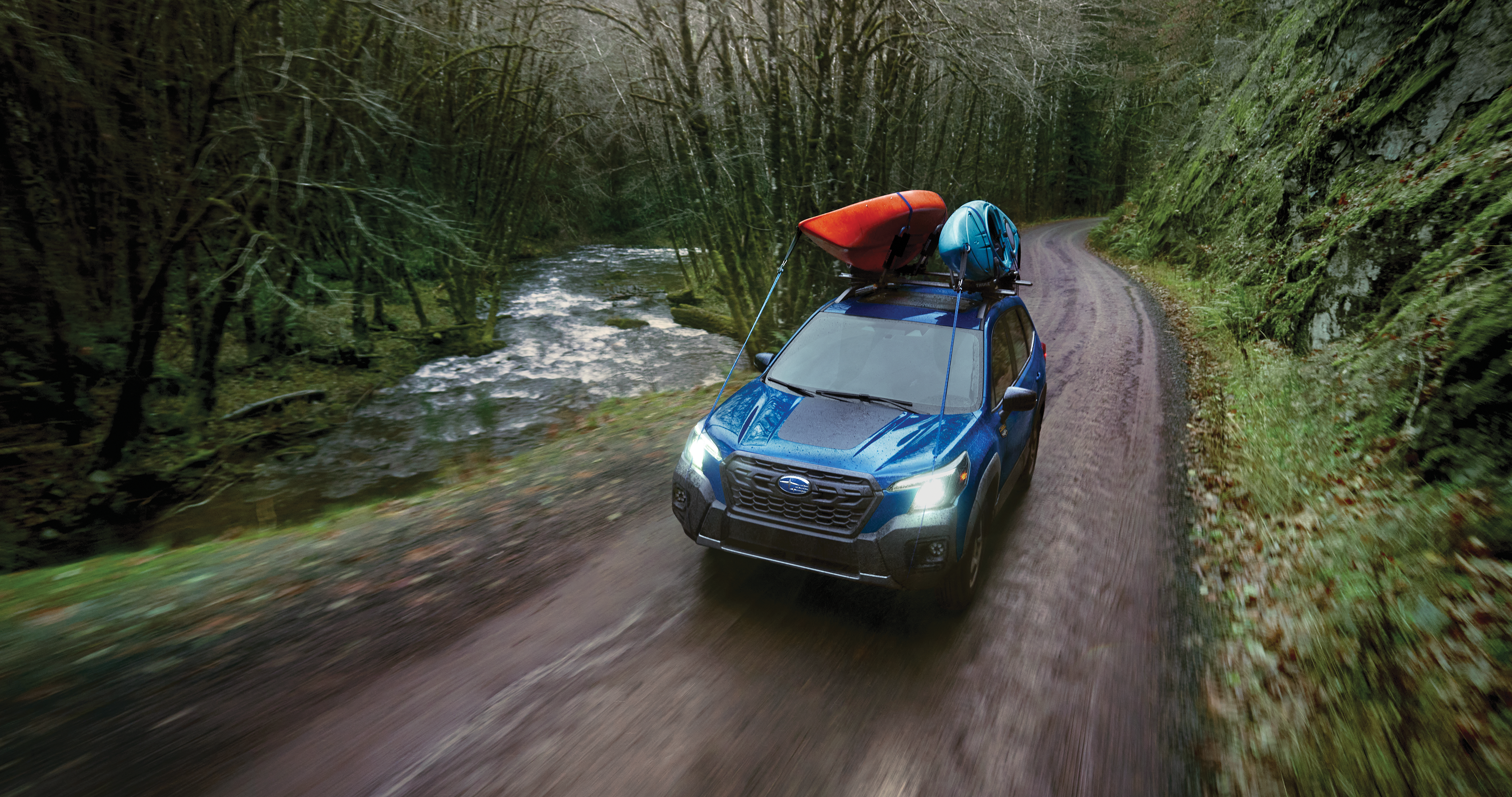 A Subaru Forester carrying two kayaks on its roof drives along a stream in the woods
