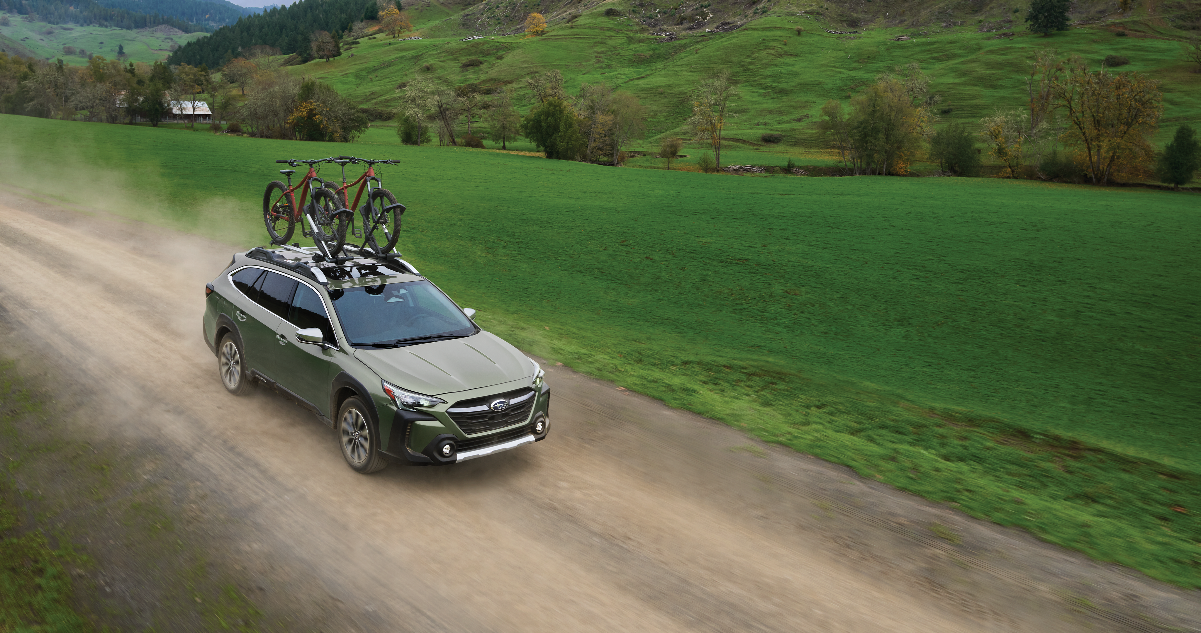 Un todoterreno Subaru Outback verde con dos bicicletas en la baca circula por un camino de grava.
