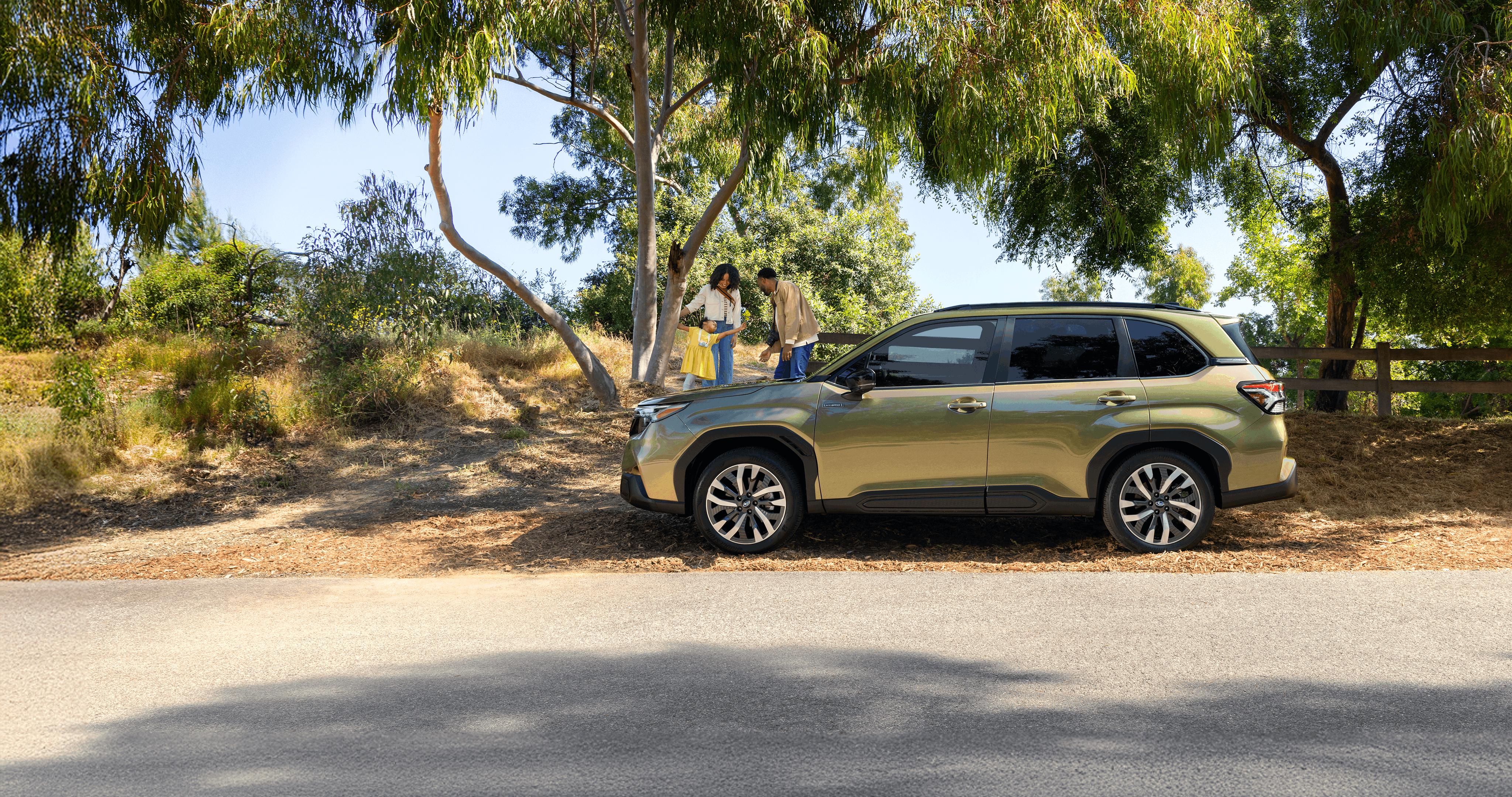 A 2026 Forester Hybrid in Autumn Green Metallic parked on the road as a family plays with their daughter in the background.