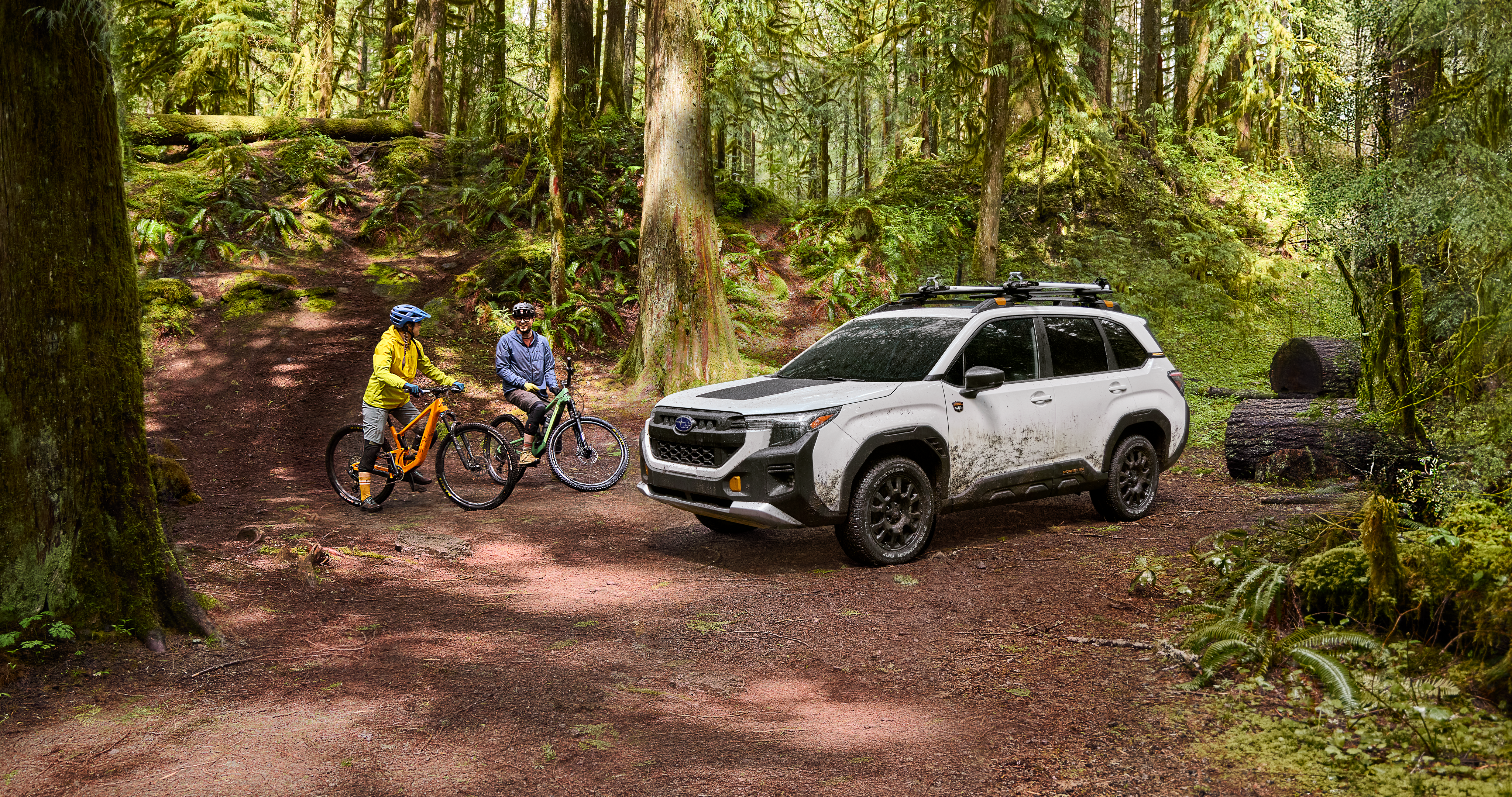 Two people on off-road bicycles next to a muddy 2026 Subaru Forester Wilderness in white in the middle of a forest.
