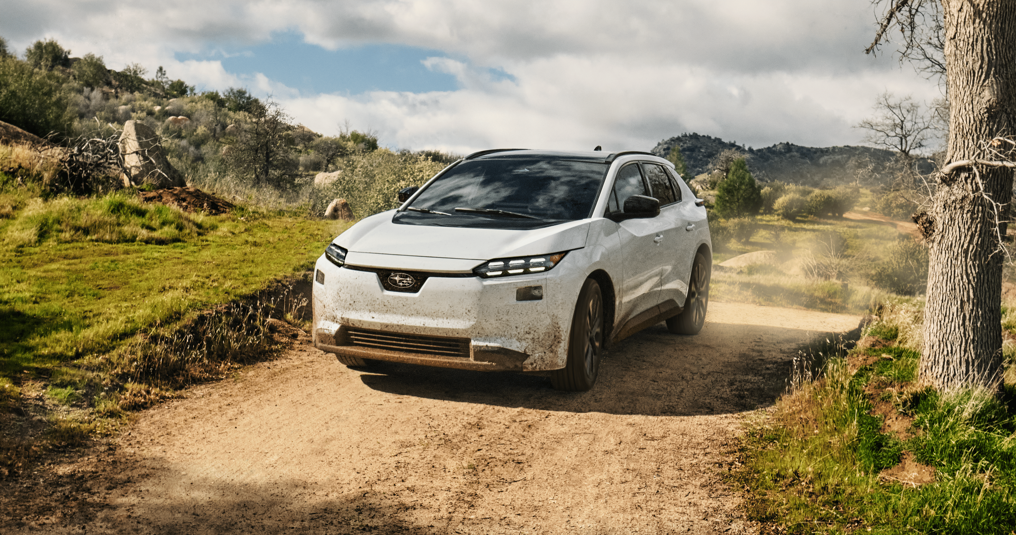 A white 2026 Subaru Solterra electric SUV drives on a dirt road in arid hills, kicking up dust behind.