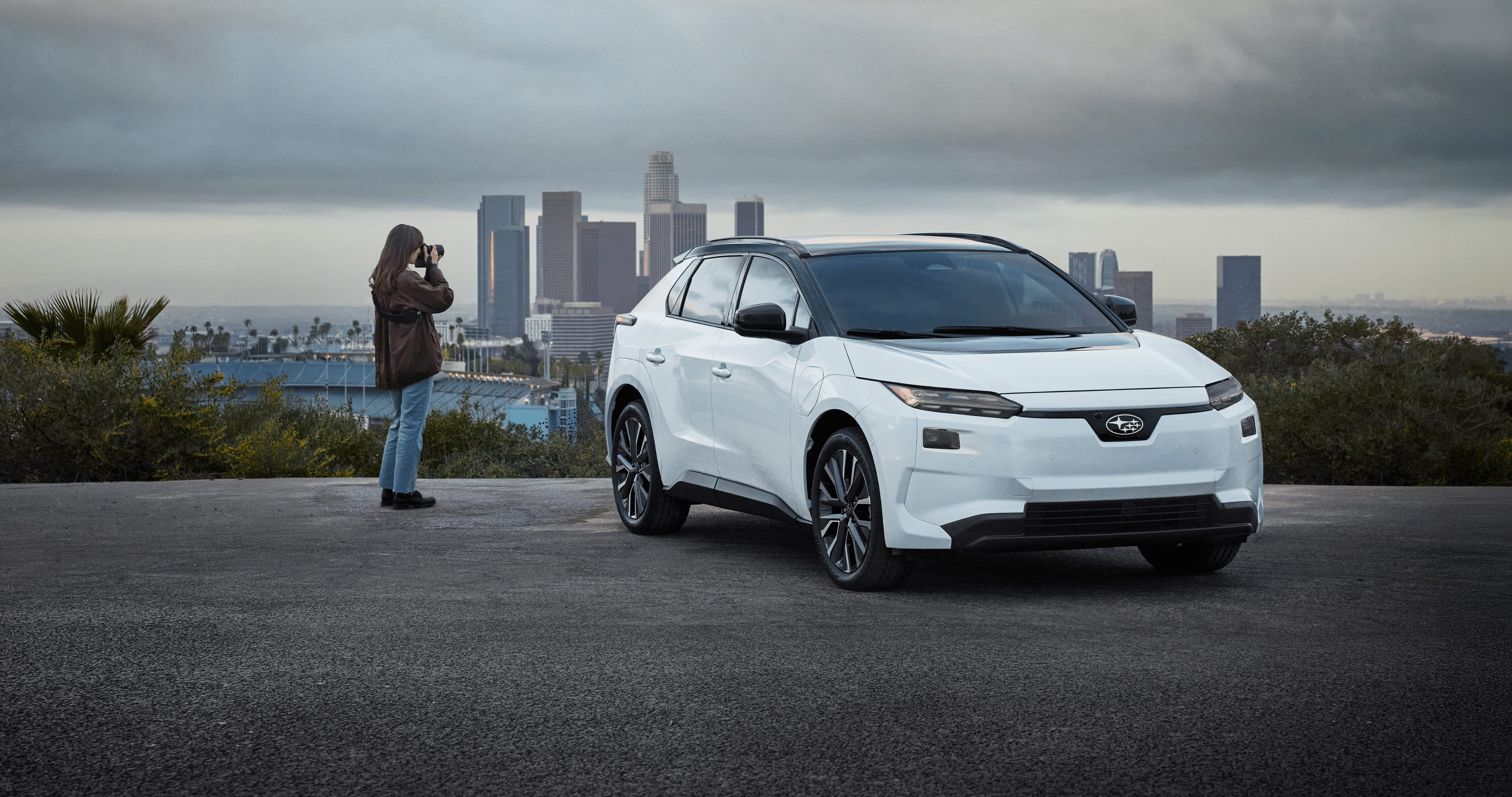 A white 2026 Subaru Solterra electric SUV drives on a dirt road in arid hills, kicking up dust behind.