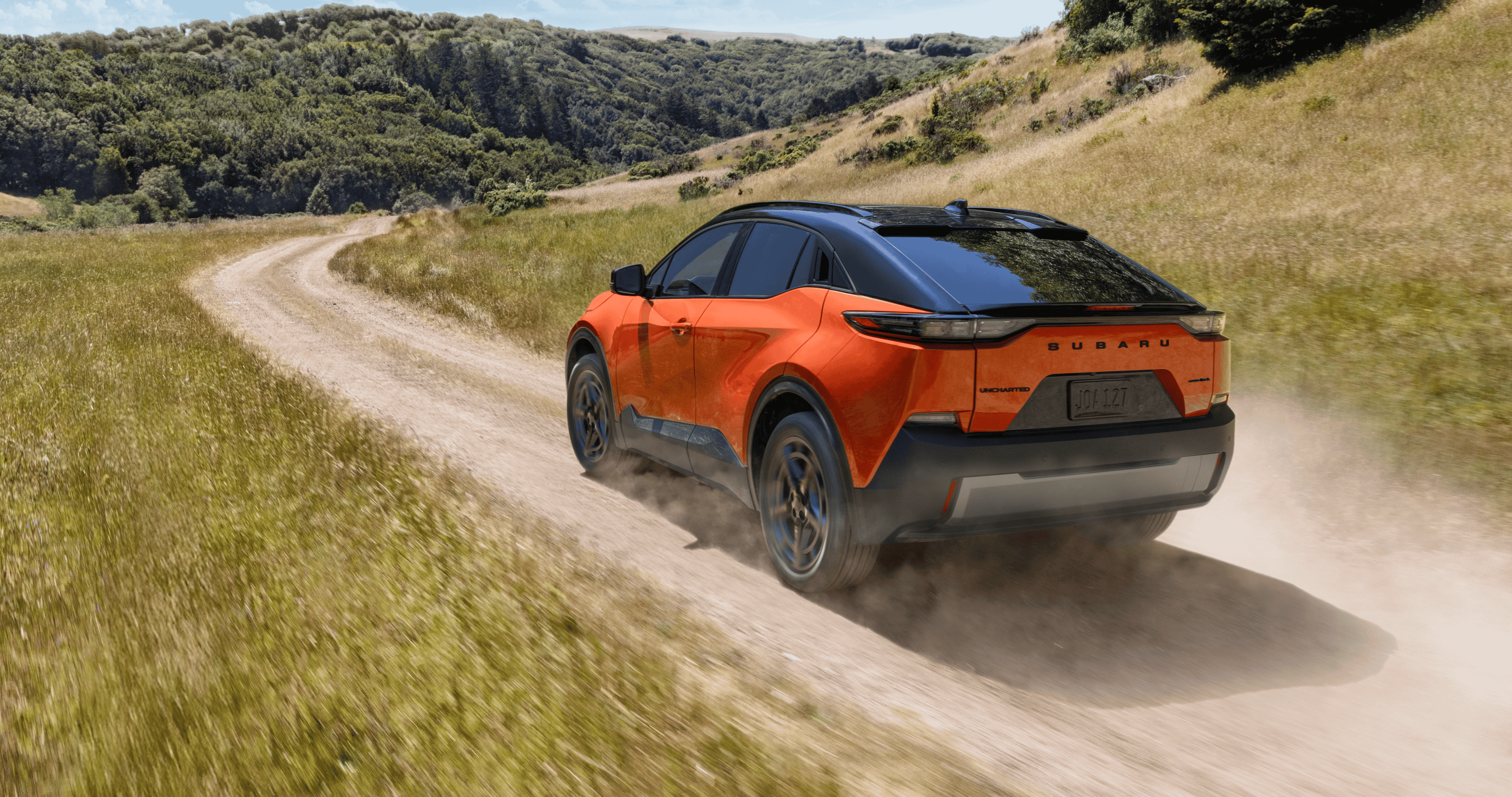 A static image of an orange 2026 Subaru Uncharted driving on a winding dirt road through wide, grassy hills.