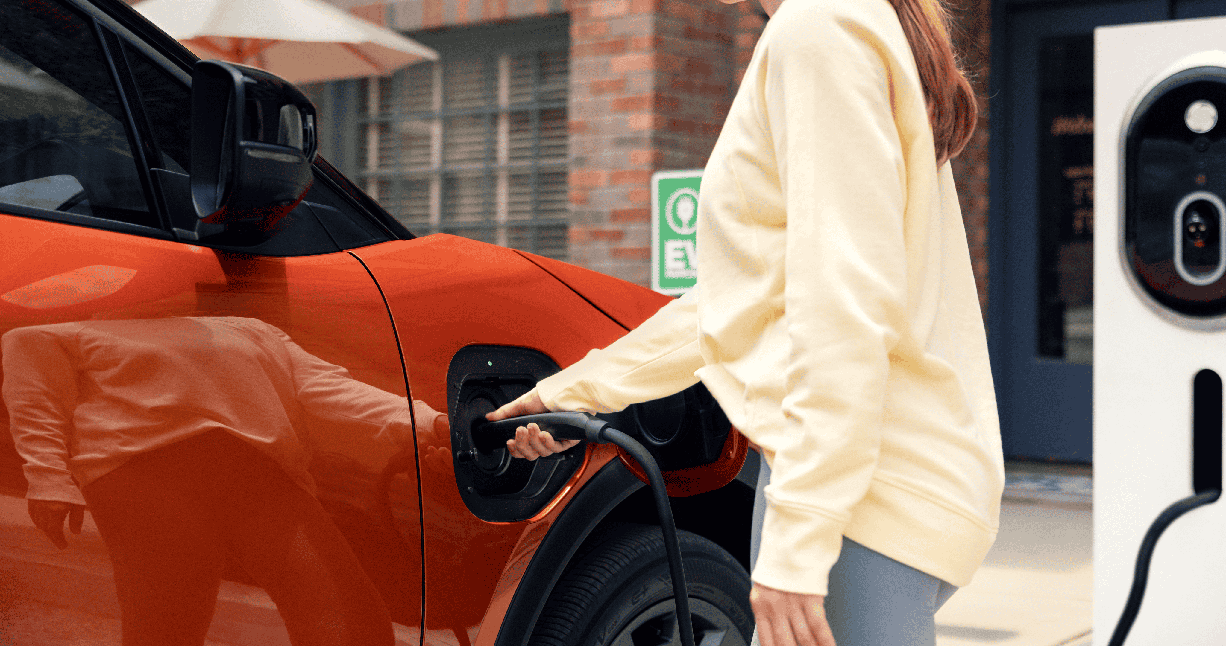 A static close-up image of a person plugging a charging cable into the front driver‑side charging port of an orange Subaru Uncharted.