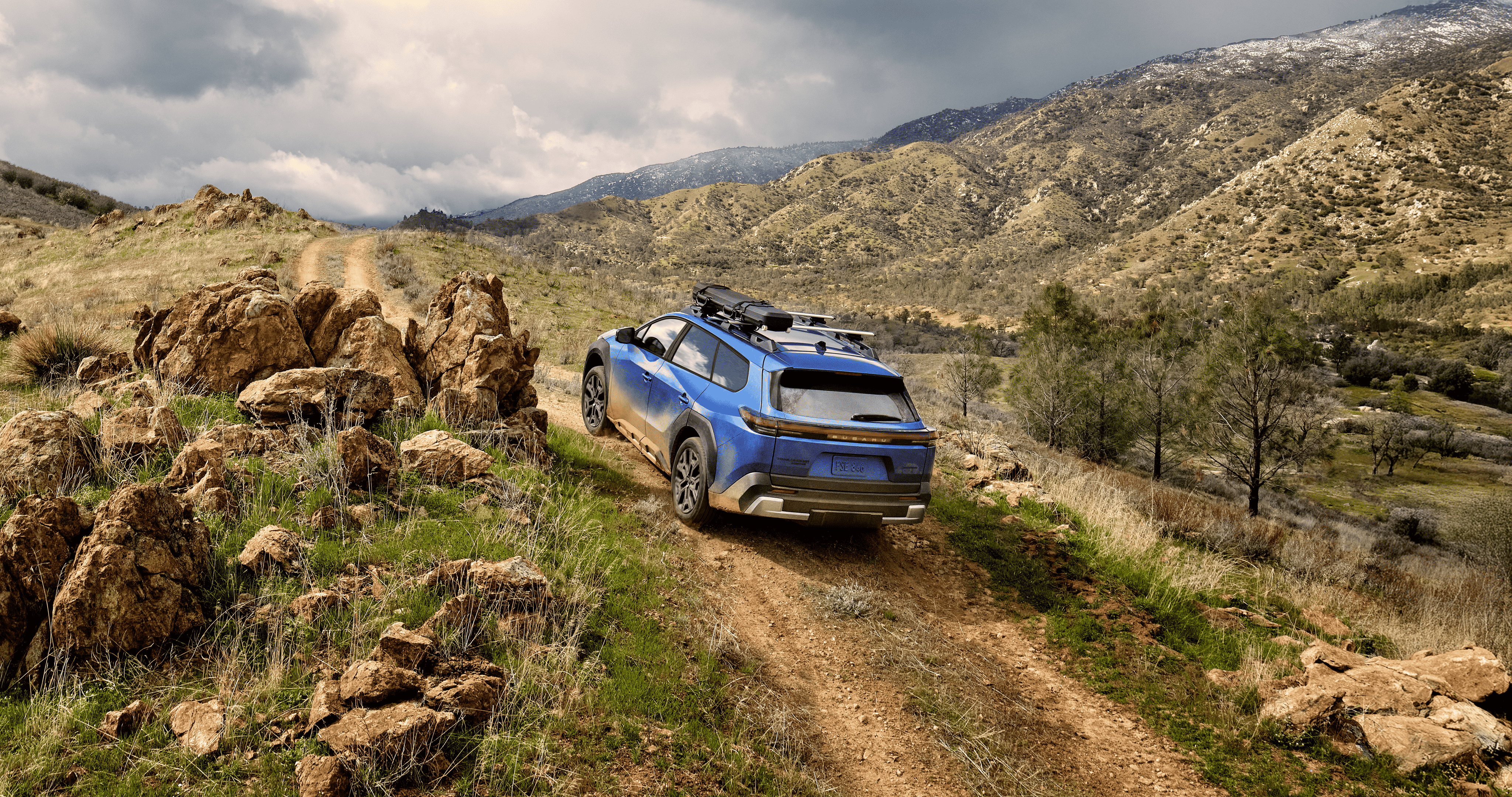 A static image of a blue 2027 Subaru Trailseeker driving on a narrow dirt trail that winds through rocky, grassy hills.