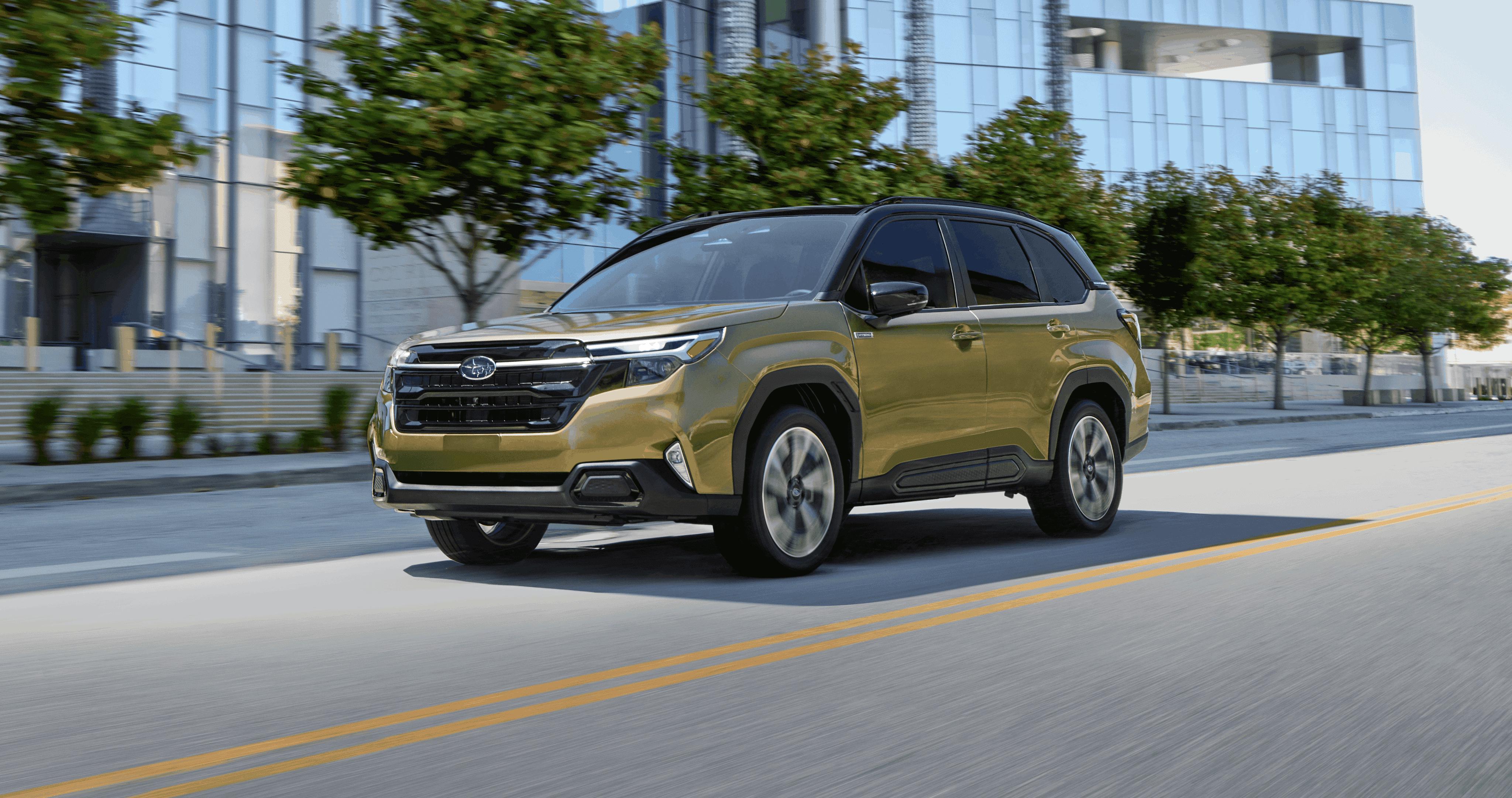 A gold Subaru Forester Hybrid drives down a city street lined with trees and modern glass buildings on a bright day.