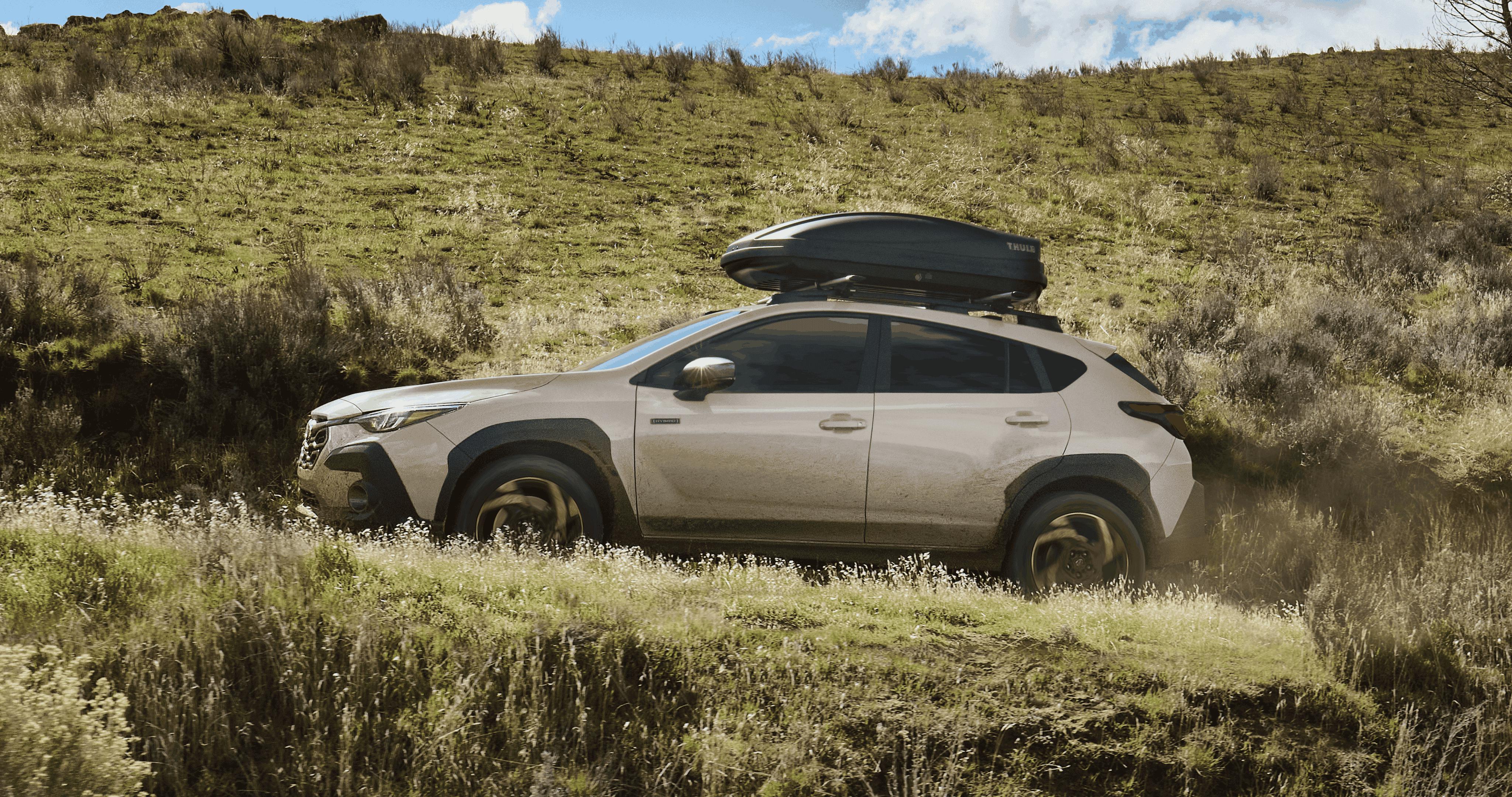 A beige Subaru Crosstrek Hybrid drives off‑road through a grassy, uneven hillside with a rooftop cargo box attached under a bright, partly cloudy sky.