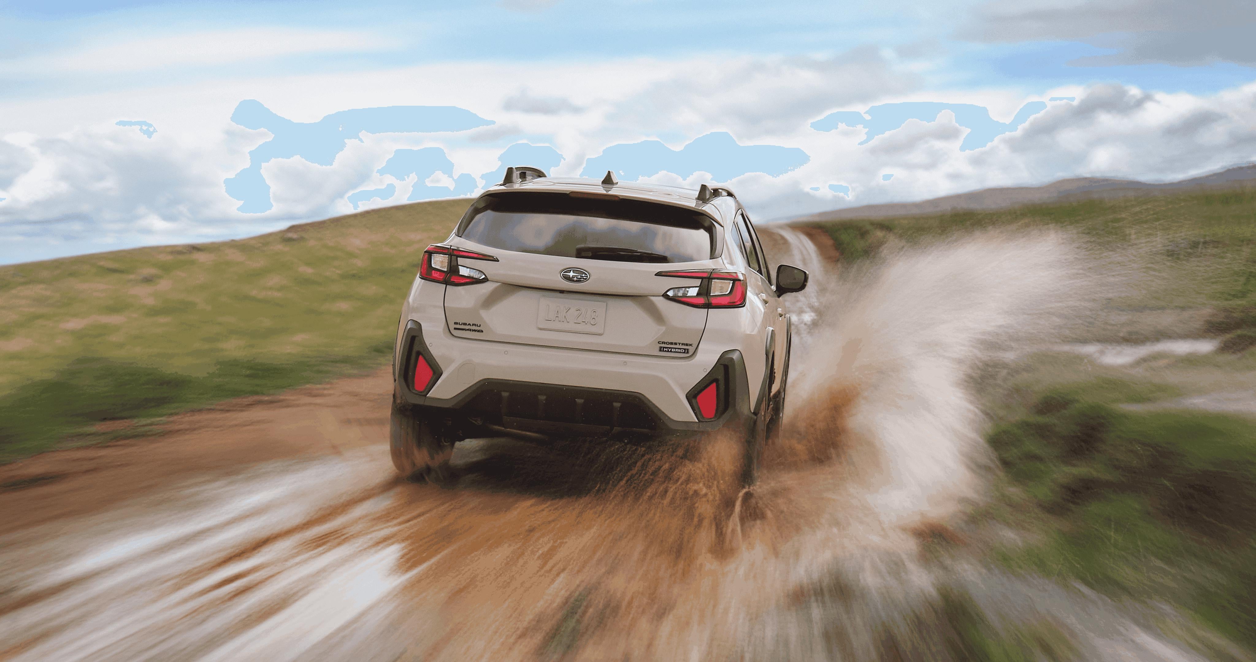 A Subaru Crosstrek Hybrid driving at speed down a muddy trail, kicking up water and mud as it moves through an open landscape under a partly cloudy sky.