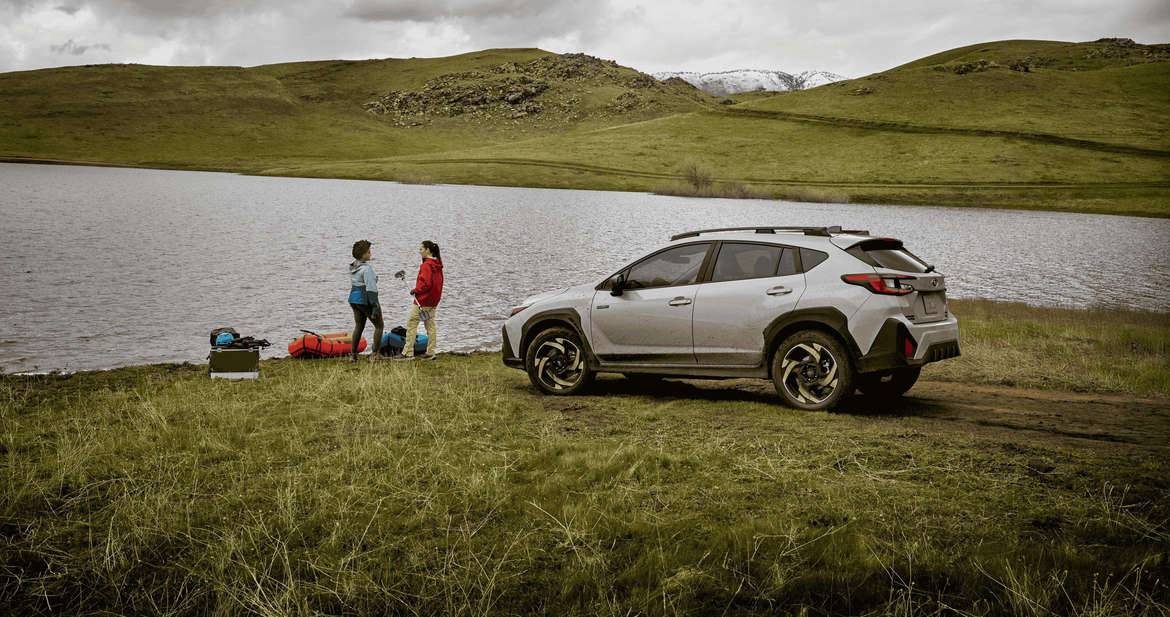 A Subaru Crosstrek Hybrid parked beside a lakeshore on a grassy field, with two people standing near camping gear by the water under a cloudy sky and rolling green hills.