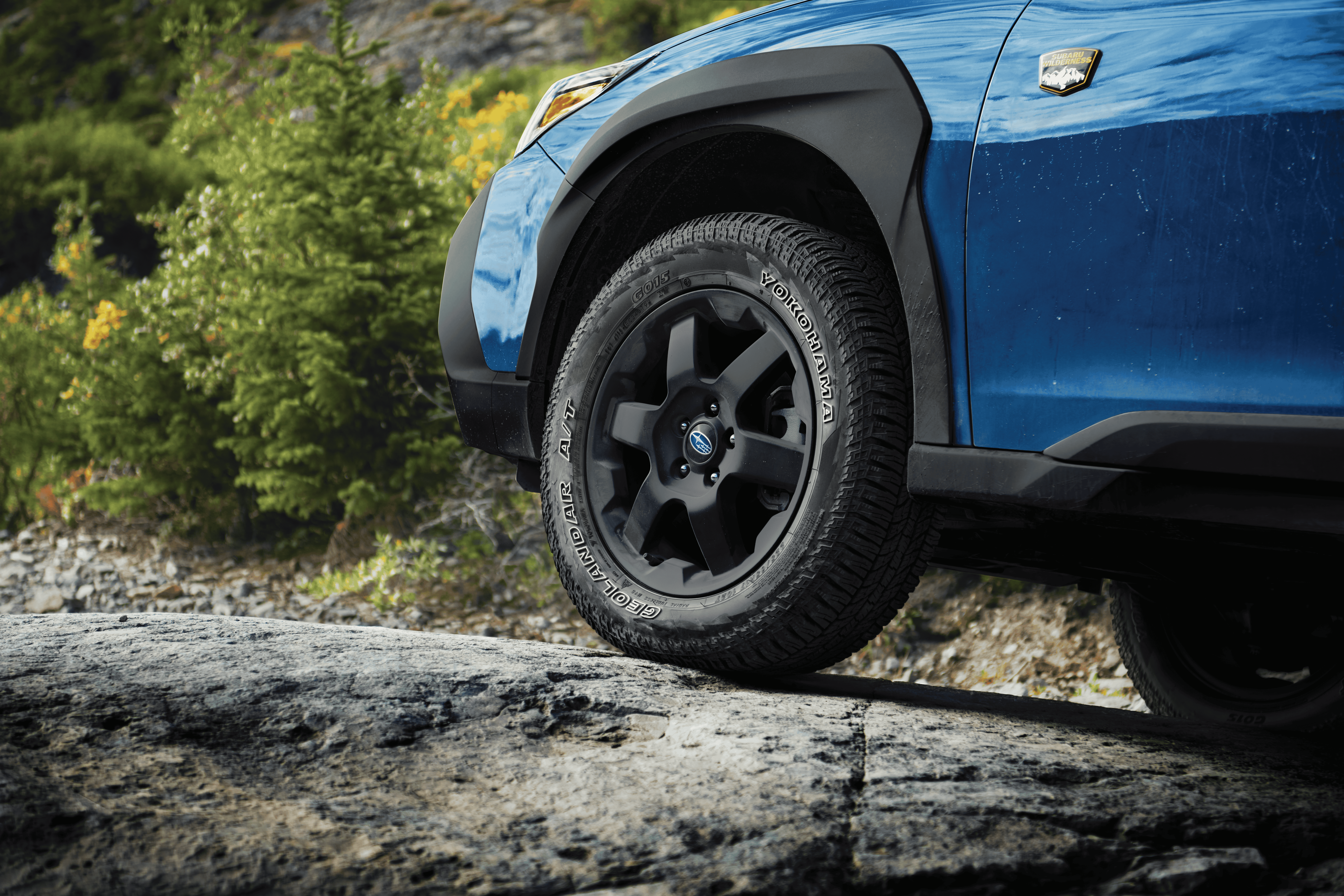 A close-up static image of the front wheel and lower side panel of a blue Subaru Wilderness model parked on uneven rocky terrain