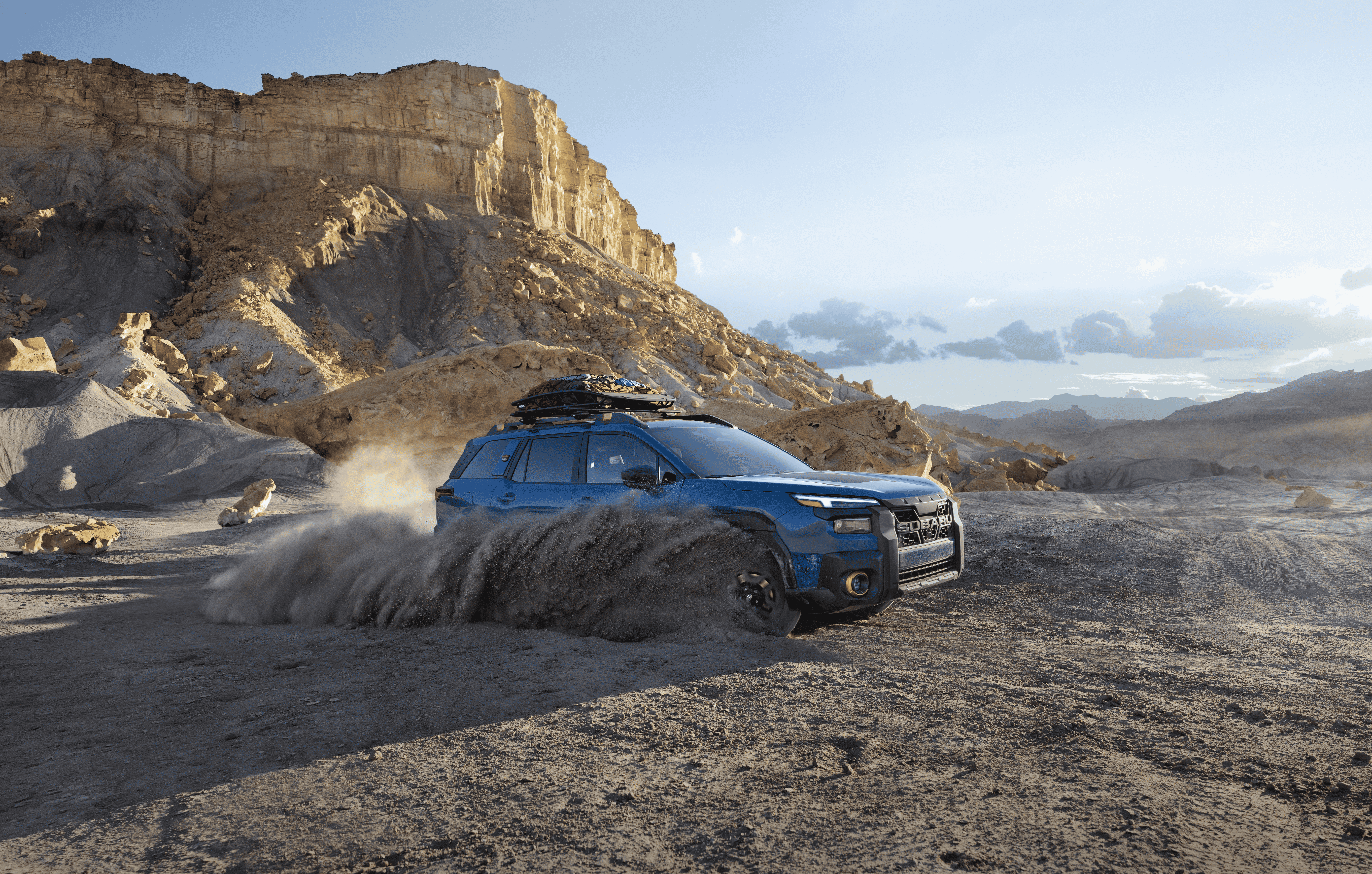 A static image of a blue 2026 Subaru Outback Wilderness driving through loose dirt in a rocky desert landscape.