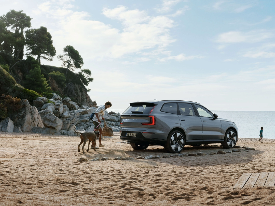 Rear-view of a Volvo EX90 fully electric car on the beach with a man, a dog, and two children running to the ocean.